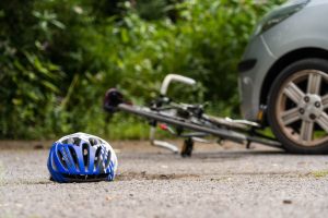 This image depicts a blue and white bicycle helmet lying on the ground in the foreground, while a bicycle is seen knocked over behind it near a car's wheel. The scene suggests a recent bike accident or collision involving the vehicle and the cyclist. The background shows greenery, and the focus is on the helmet, with the rest of the scene slightly blurred, emphasizing the possible aftermath of the event.