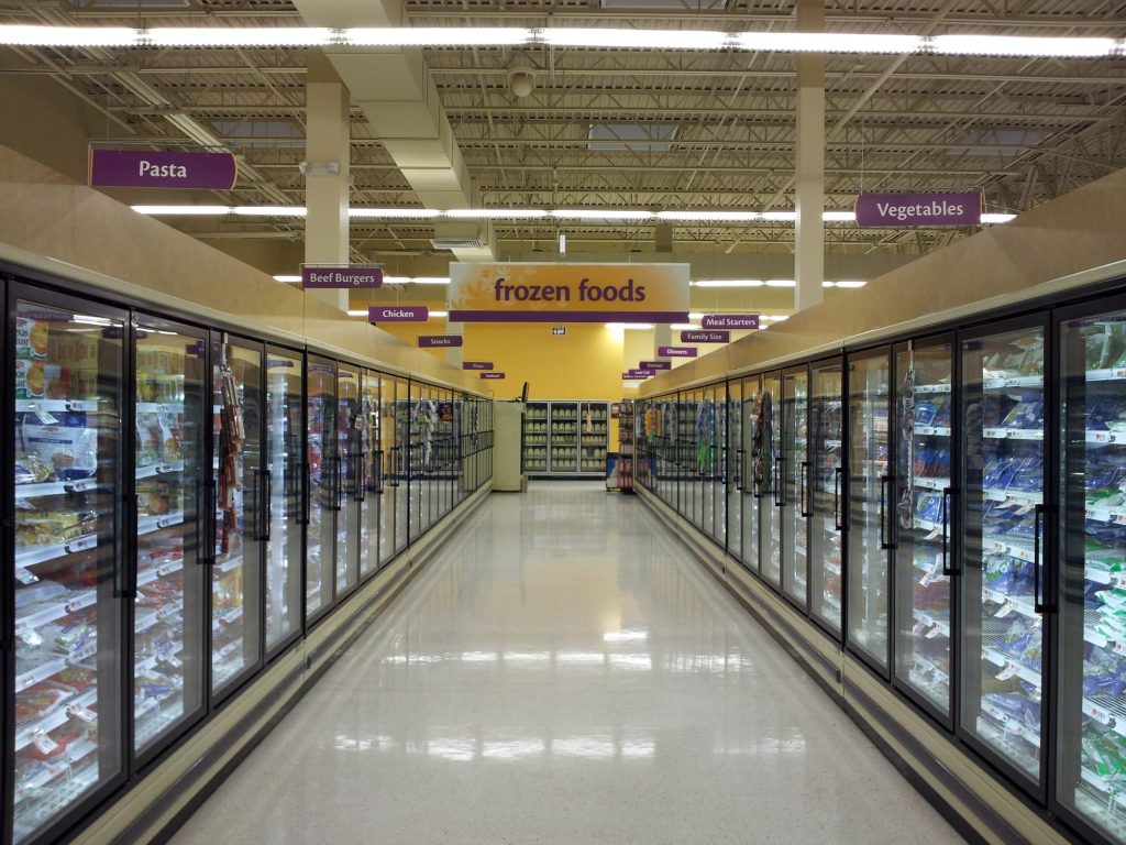 This image shows a clean, well-lit grocery store aisle dedicated to frozen foods. Both sides of the aisle feature glass-door freezers containing various items. Signs hanging from the ceiling label the sections, including "Pasta," "Chicken," "Beef Burgers," "Vegetables," and "Meal Starters." The floor is bright and reflective, and the arrangement is orderly, with products clearly visible through the freezer doors. The store appears spacious, with a focus on organization and accessibility.