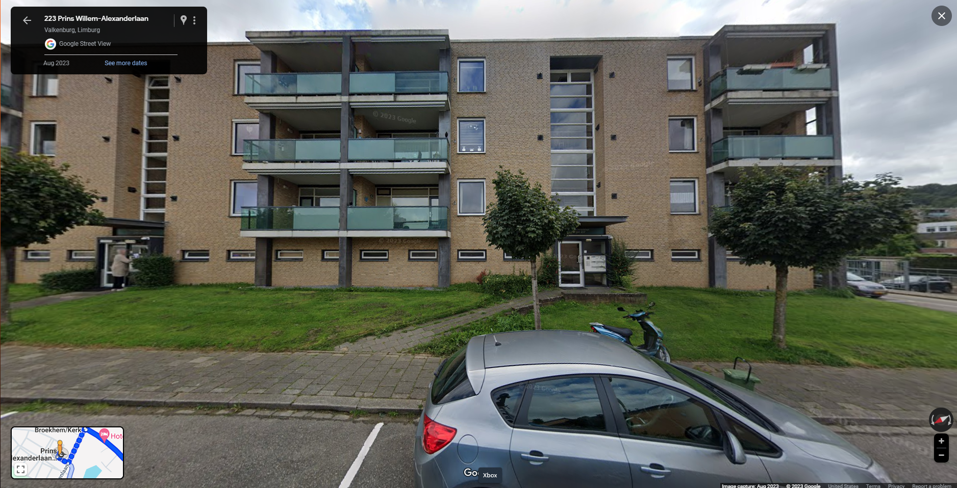 This image shows a Google Street View capture of a three-story brick apartment building at 223 Prins Willem-Alexanderlaan in Valkenburg, Limburg, Netherlands. The building features balconies with glass railings and green lawns in front. A person can be seen entering one of the entrances on the left side. In the foreground, a gray car is parked on the street near a small tree and a moped. The image includes a map inset in the bottom-left corner, displaying the location and surrounding streets. The timestamp in the top-left corner shows the image was captured in August 2023.