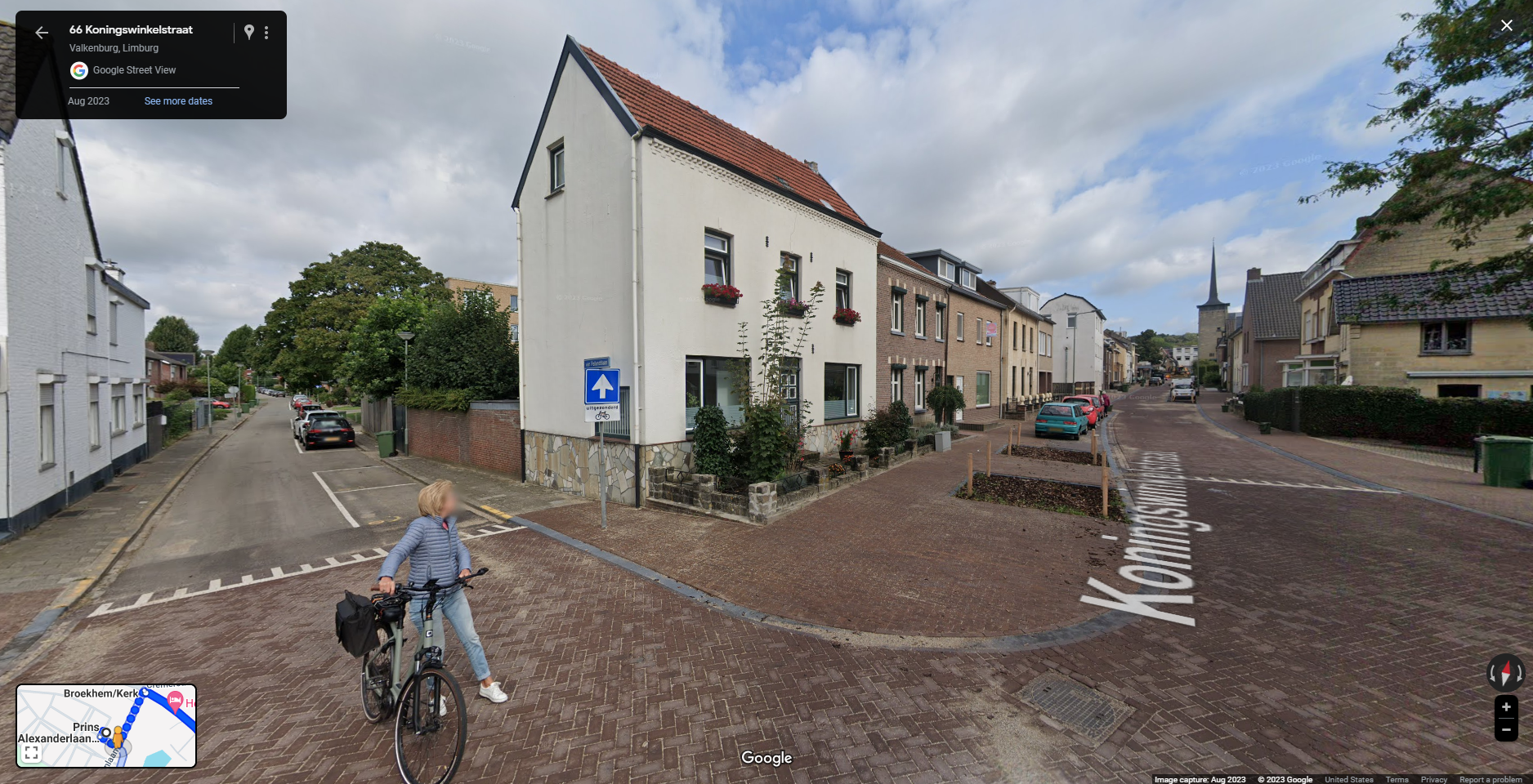 This image shows a Google Street View capture of 66 Koningswinkelstraat in Valkenburg, Limburg, Netherlands taken in August 2023. It depicts a quiet residential street with brick houses, flower boxes on some windows, and a cyclist standing near the intersection, facing away from the camera. The street is paved with bricks, and there are cars parked along both sides of the road. In the distance, a church steeple is visible. The inset map in the lower-left corner shows the location and nearby roads. The overall setting is peaceful, capturing a typical street scene in a small European town.