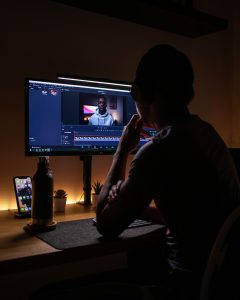 This image shows a person sitting at a desk, editing a video on a large monitor. The individual is wearing headphones and appears to be focused on the screen, which displays video editing software with a clip of someone speaking. The desk is neatly arranged, featuring a water bottle, a smartphone on a stand, and small decorative plants, all softly lit by warm ambient lighting. The scene conveys a calm, focused atmosphere typical of a creative workspace.