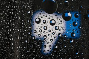 This image shows water droplets on a glass surface, with each droplet reflecting small "thumbs up" icons, similar to the Facebook "Like" symbol. The background appears to be blurred, showing a larger thumbs-up icon in blue and white, creating an interesting pattern as the reflections multiply within the droplets. The image is a close-up, highlighting the water droplets and their reflections in sharp detail. The overall effect gives a creative and visually striking representation of social media engagement.